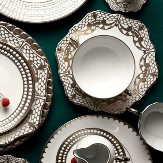 L'Objet Aegean Filet Platinum Dinnerware—plates, bowls, and cups—features elegant white pieces with platinum accents, artfully arranged with silverware on a dark surface to highlight their luxurious design.