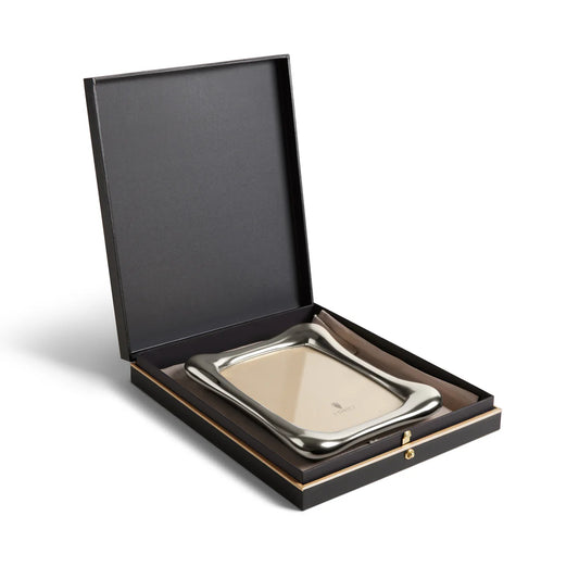 The Anton Frame Platinum by L'Objet is a platinum-plated tray with wavy edges, displayed in a black hinged gift box lined with brown fabric and secured by a gold clasp, embodying the refined elegance of a luxury photo frame.