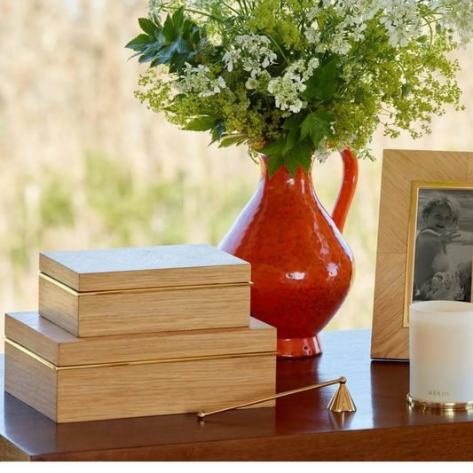 A wooden table displays an Aerin Marcello Box Oak stacked with an Art Deco wood box, a gold candle snuffer, a white candle, a framed black-and-white photo, and a red vase filled with white and green flowers.