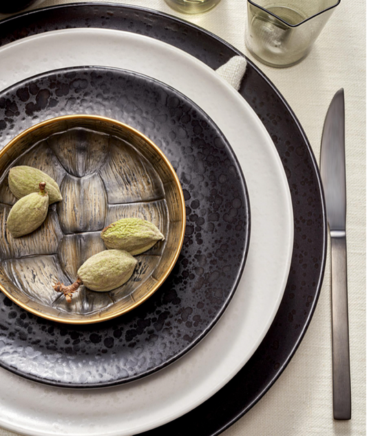 A rustic place setting featuring three L'Objet Alchimie Dinnerware White plates, a small metallic bowl filled with green almonds, and a dark gray knife.