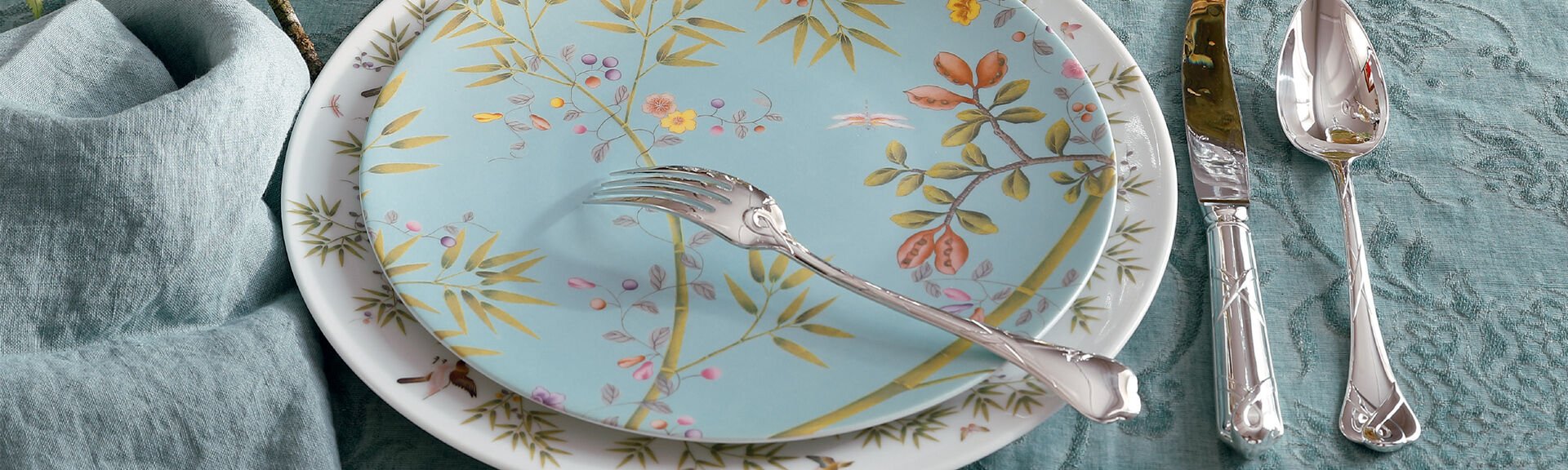 A close-up of a table setting with two decorative plates featuring floral patterns, a silver fork resting on top, and a knife and spoon on a blue tablecloth.