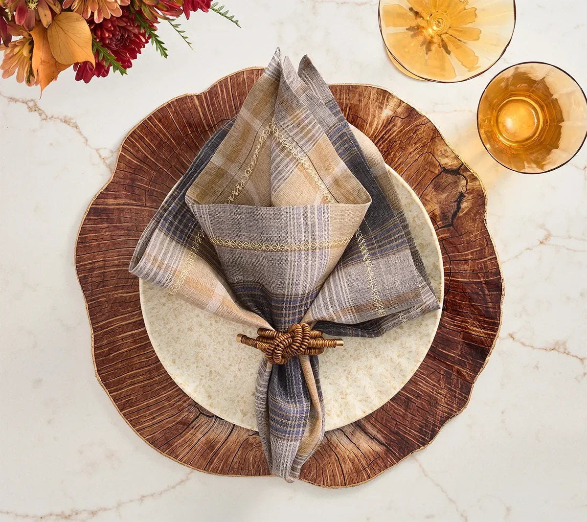 A rustic table setting with a wood-look charger, beige plate, plaid napkin in a woven napkin ring, and two amber glasses on a marble surface. Autumn flowers are partially visible in the corner.