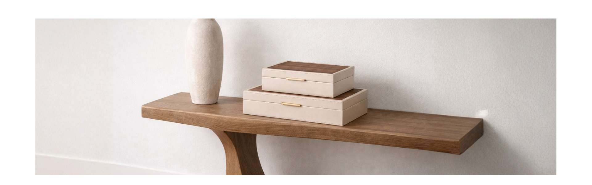 A minimalist wooden console table holds a tall, textured beige vase and two stacked, rectangular beige boxes with wooden lids against a light-colored wall.