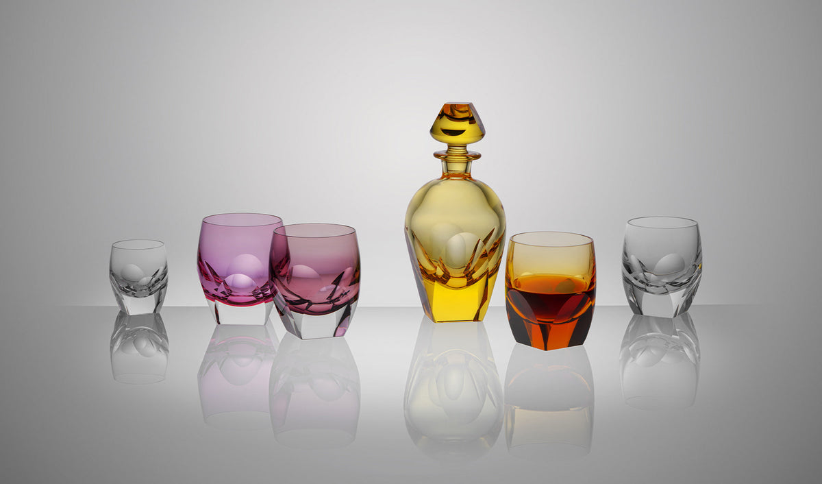 Six crystal glasses and one decanter in pink, yellow, and clear glass are arranged in a row on a reflective surface, with soft lighting and a neutral gray background.