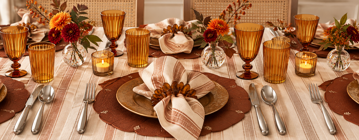A fall-themed table setting with amber glasses, striped napkins, orange flowers in small vases, candles, and brown placemats arranged neatly for a festive meal.