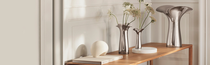 Minimalist wooden table with decorative items: two silver vases with white flowers, a large silver sculptural vase, a round white object, a white tray, and two stacked books against a white paneled wall.