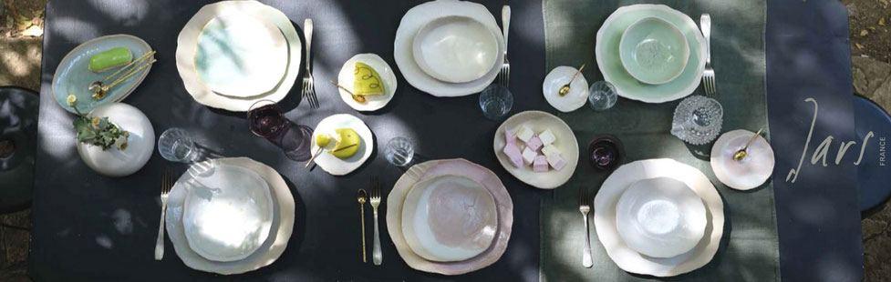 A table set for six with white plates, glassware, gold utensils, and small dishes of food, arranged on a dark tablecloth with dappled sunlight.