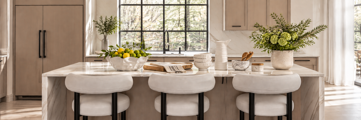 Modern kitchen with a marble island, three white cushioned stools, a bowl of lemons, a vase of green flowers, and large windows letting in natural light.