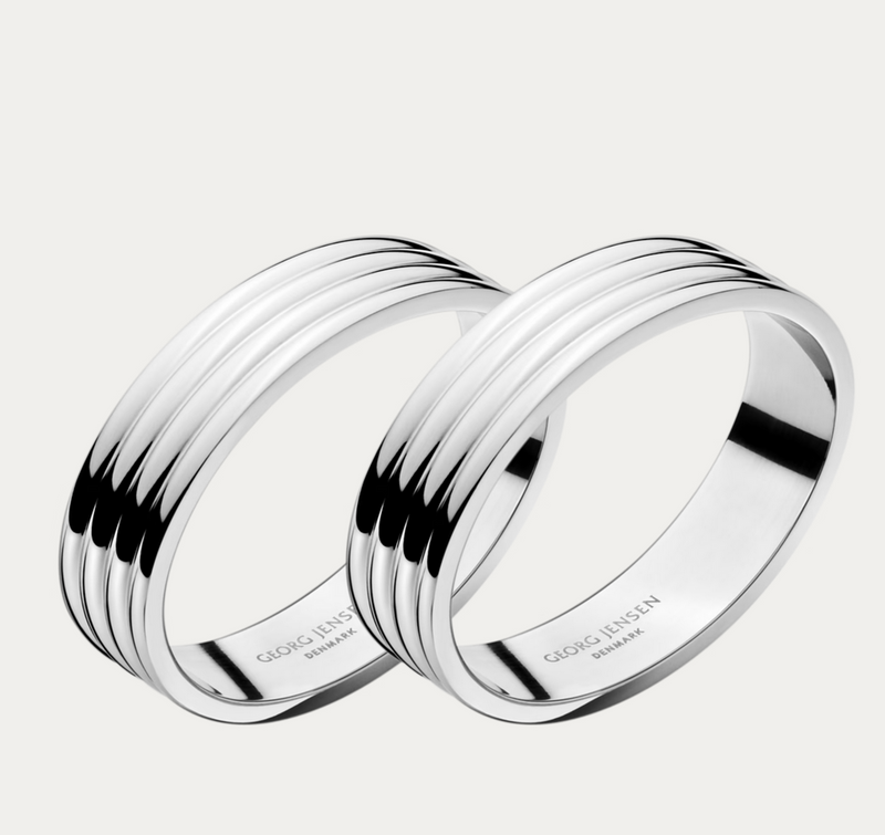 The Bernadotte Napkin Ring Set of 2 by Georg Jensen features silver-toned stainless steel rings with a signature triple-groove design, shown overlapping on a plain white background. Each band is engraved with the Georg Jensen name on the inside.