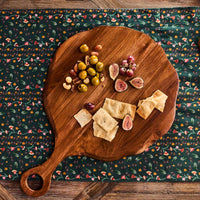 The Juliska Puro Round Serving Board Teak showcases an inviting arrangement of green olives, nuts, figs, grapes, and pieces of flatbread atop a floral-patterned tablecloth—perfect for entertaining with a beautifully presented charcuterie board.