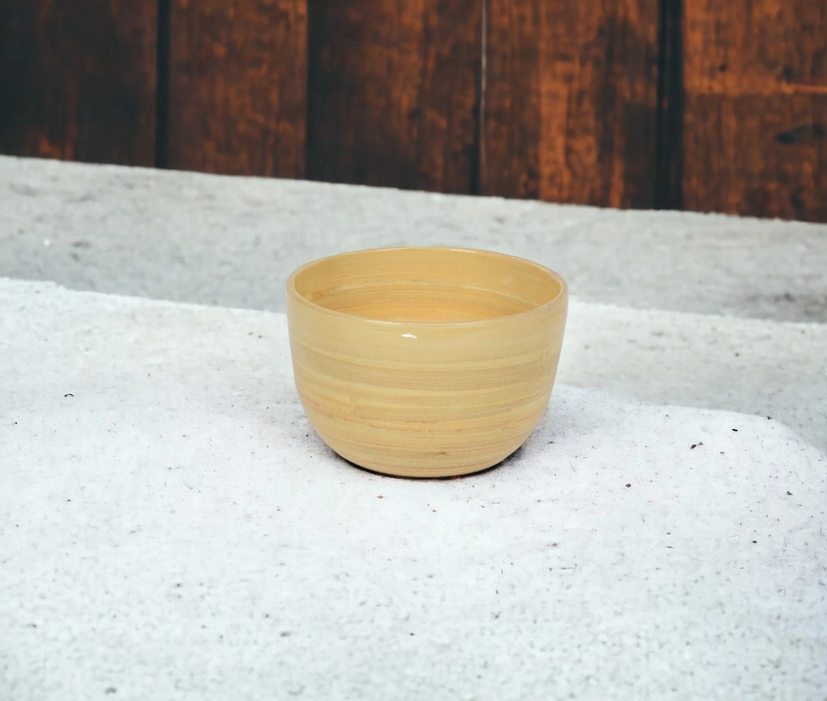 The Albert L Bamboo Classic Serving Bowl Small in Natural rests on a textured white surface with a wooden wall behind it.