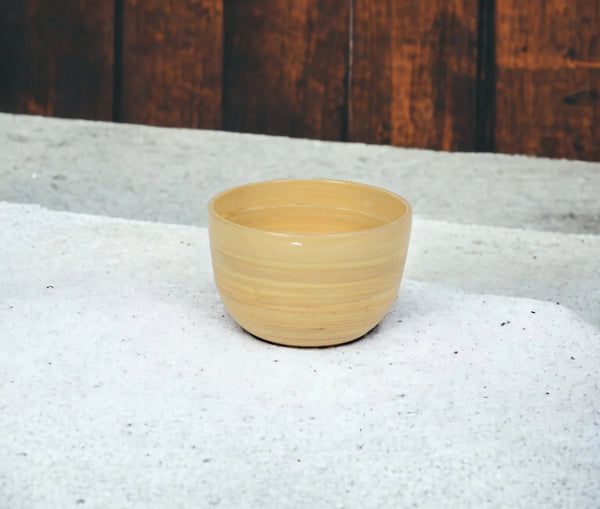 The Albert L Bamboo Classic Serving Bowl Small in Natural rests on a textured white surface with a wooden wall behind it.