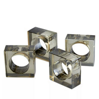 Four transparent square napkin rings from Tizo’s Crystal Cube Napkin Ring Smoke Set of 4 are arranged on a white surface. These modern geometric rings feature circular openings and a smoky grey color that reflects light, creating an elegant, contemporary look reminiscent of smoke-inspired designs.