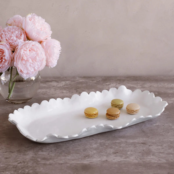 The Beatriz Ball Vida Colette Platter White - Large, featuring wavy edges, displays four colorful macarons on a gray surface beside a glass vase of soft pink peonies.