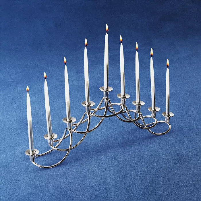 The Georg Jensen Harmony Menorah, featuring a modern silver design and nine lit white Hanukkah candles, is displayed on a blue surface with a blue background.
