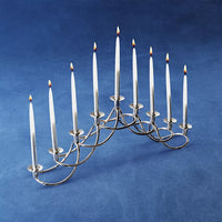 The Georg Jensen Harmony Menorah, featuring a modern silver design and nine lit white Hanukkah candles, is displayed on a blue surface with a blue background.