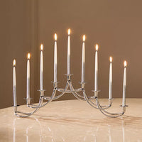 A Georg Jensen Harmony Menorah with nine lit white Hanukkah candles sits on a marble surface against a beige background.