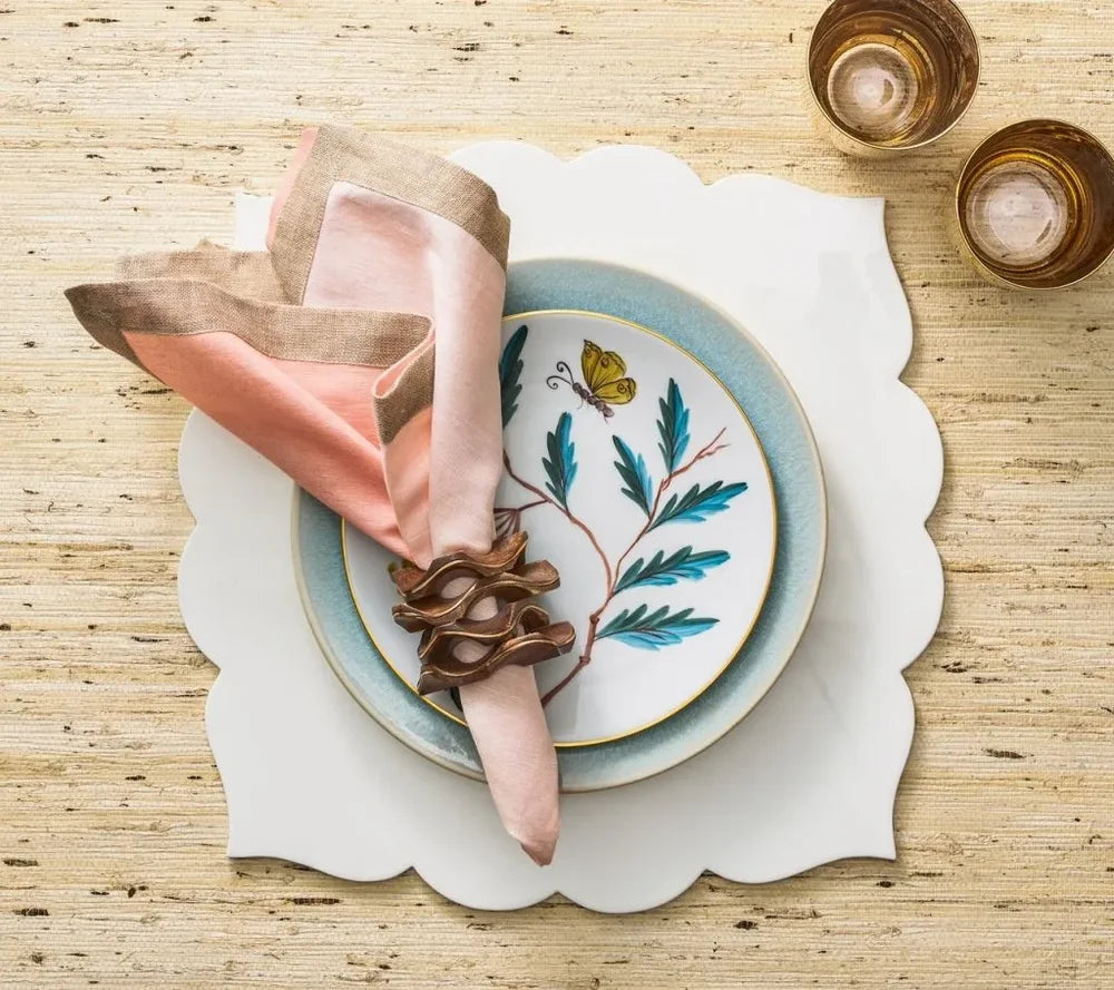 A decorative plate featuring a butterfly and leaf motif, adorned with a pink and beige napkin in a wooden holder and layered with additional plates, is complemented by two amber glasses—all beautifully arranged on the Guzzini Fez Placemat (Set of 4) for an elegant touch of style.