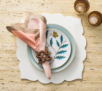 A decorative plate featuring a butterfly and leaf motif, adorned with a pink and beige napkin in a wooden holder and layered with additional plates, is complemented by two amber glasses—all beautifully arranged on the Guzzini Fez Placemat (Set of 4) for an elegant touch of style.