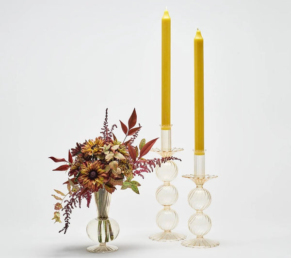 Kim Seybert’s Bella Candlestick Holder Set of 2, made from clear hand-blown glass and featuring tall yellow candles, stands beside a small glass vase filled with fall-colored flowers and foliage, all set against a plain white background.