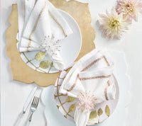 Two elegant place settings featuring Guzzini’s Fez Placemats in a chic champagne scallop design, white plates adorned with gold leaf patterns, white napkins embroidered with gold, clear and pink napkin rings, and two dahlias in the top right corner. The fork and knife are placed to the left of each setting.