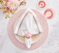 A cream plate holds a white napkin with decorative stitching, secured by the Kim Seybert Sea Stone Napkin Ring Blush Set of 4, atop a textured pink placemat. Two pink drinks and a vase of pink flowers sit nearby on a marble surface.