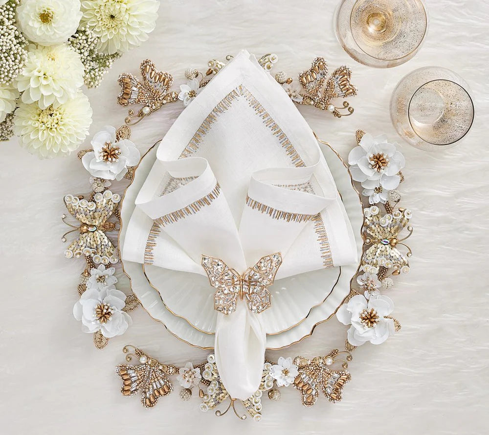 An elegant table setting features white florals, gold accents, a scalloped-edge plate, and a white napkin with a Kim Seybert Papillon Butterfly Napkin Ring (Boxed Set of 4), plus two glasses on a textured white surface—perfect for luxury decor.
