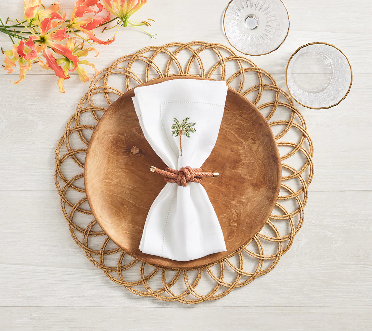 A Kim Seybert Alfresco Placemat in Natural sets the scene, topped with a wooden plate, white embroidered napkin tied with brown cord; glassware and orange flowers complete this inviting outdoor dining table.
