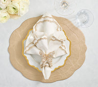 Create a luxurious table setting with layered beige and white plates, an embroidered napkin held by a Kim Seybert Papillon Butterfly Napkin Ring, clear glasses on a white tablecloth, and a bouquet of white flowers.