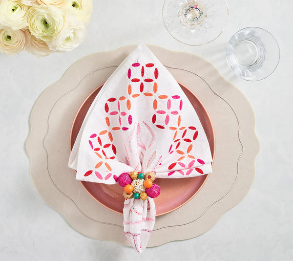 A white napkin with a pink and orange geometric pattern sits in a beaded ring on a pink plate and scalloped cream charger, all atop the Kim Seybert Tailored Placemat Set of 4 in Ivory, beside a glass and fresh white flowers.