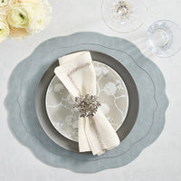 Elegant table setting featuring the Kim Seybert Tailored Placemat Set of 4 in Steel Grey, paired with a silver charger, patterned plate, cream napkin with ring, crystal wine and water glasses, and white ranunculus flowers in the upper left.