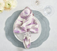 A white plate sits on the Kim Seybert Tailored Placemat in Steel Grey, topped with a white and purple napkin in a glass ring. Nearby are a wine glass, water glass, and white flowers.