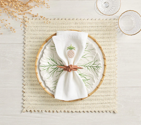 A white plate with a green leaf motif rests on a Kim Seybert Vista Reversible Placemat Set of 4 in Ivory & Beige, accented by a pineapple-embroidered napkin and brown ring. Clear glassware and dried flowers adorn the light wood table.