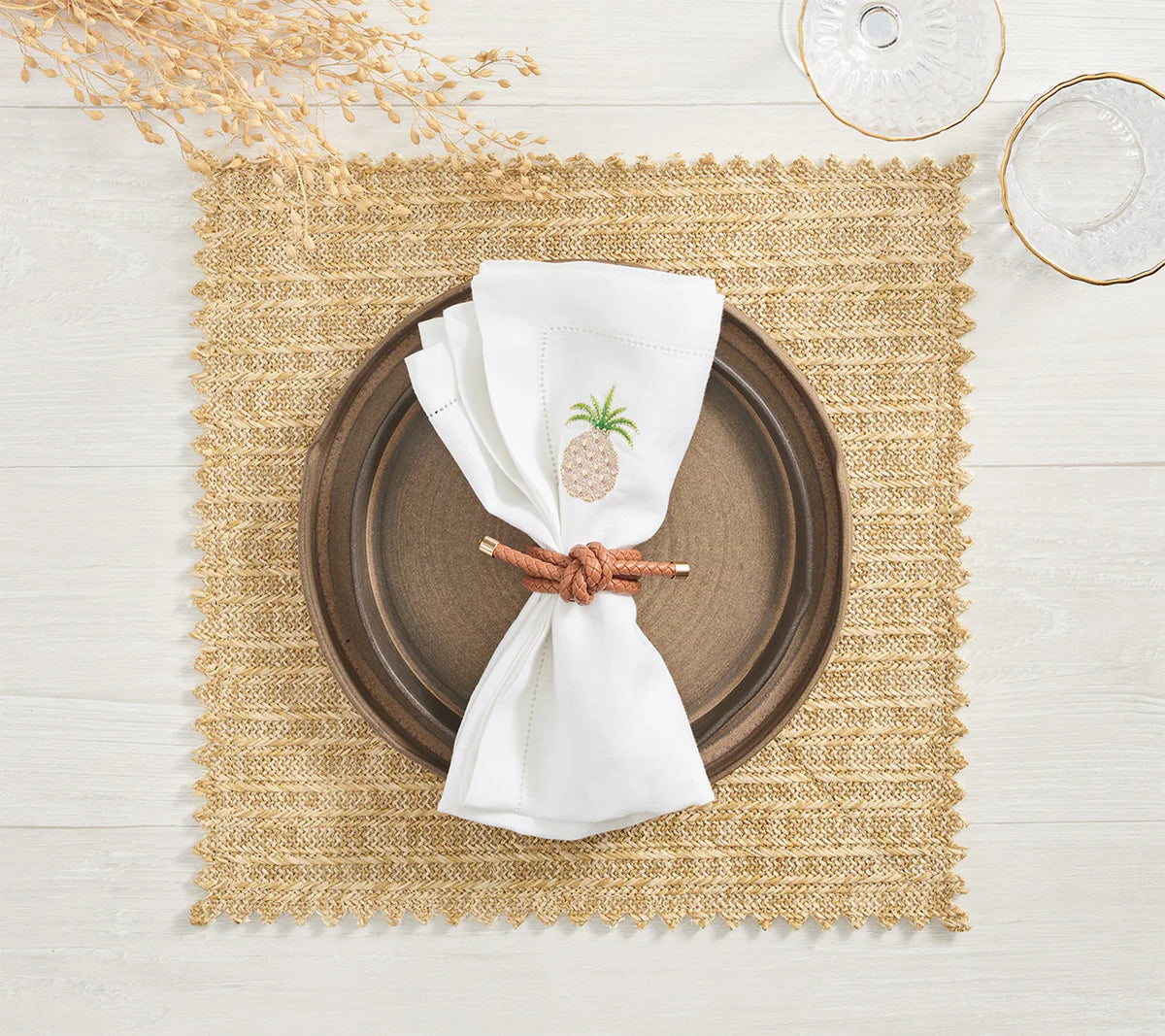 A brown plate rests on a Kim Seybert Vista Reversible Placemat in ivory & beige, with a white napkin featuring an embroidered pineapple and brown napkin ring. Two clear glasses and dried foliage sit on the light wooden table.
