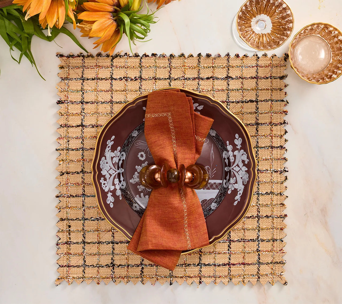 A square, brown Boucle Reversible Placemat set of 4 featuring a plate topped with a folded napkin and ring, set on a table with glassware and flowers for stylish table decor.