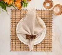 A square brown plaid Boucle Reversible Placemat set of 4 - Brown / Multi with fringed edges under a white plate, a beige linen napkin, and a gold-toned napkin ring, offering stylish table decor.
