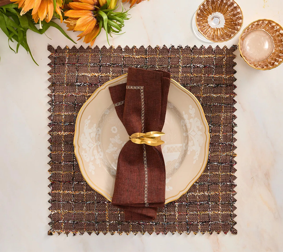 A brown and multi Boucle Reversible Placemat set of 4 featuring a square shape, brown plaid pattern, and zigzag edge, styled with a decorative plate, a folded dark napkin, and a gold leaf-shaped napkin ring.