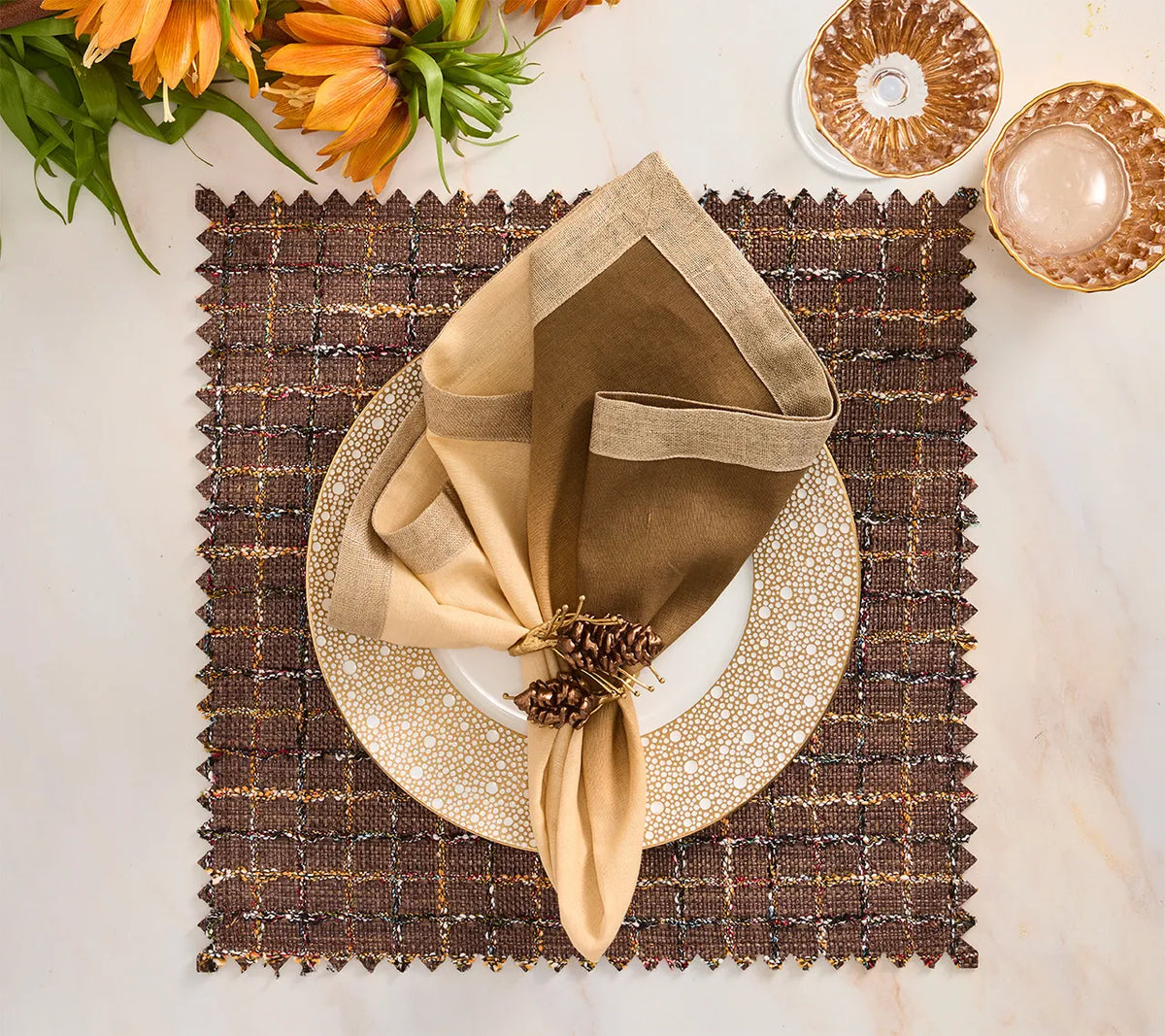 A square brown Boucle Reversible Placemat set of 4 - Brown / Multi with jagged edges, topped with a white-and-gold dotted plate and a folded cream and brown napkin held by a pinecone napkin ring.