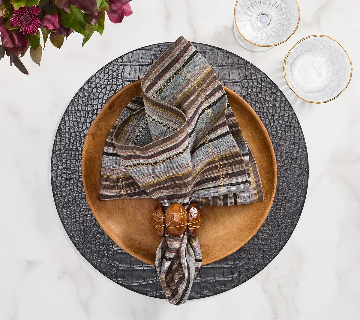 A wooden bowl holding a Windsor Napkin from the Kim Seybert Napkin Set of 4 - Multi rests on a black textured charger plate, accompanied by two clear glasses with gold rims atop a white marble surface; neutral tones and hints of purple flowers can be seen in the corner.