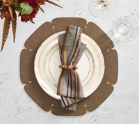 A plate with a striped Windsor Napkin from Kim Seybert and a modern gold napkin ring sits on a brown placemat, surrounded by neutral tones. Two empty glasses and a floral arrangement are nearby on a white textured surface.