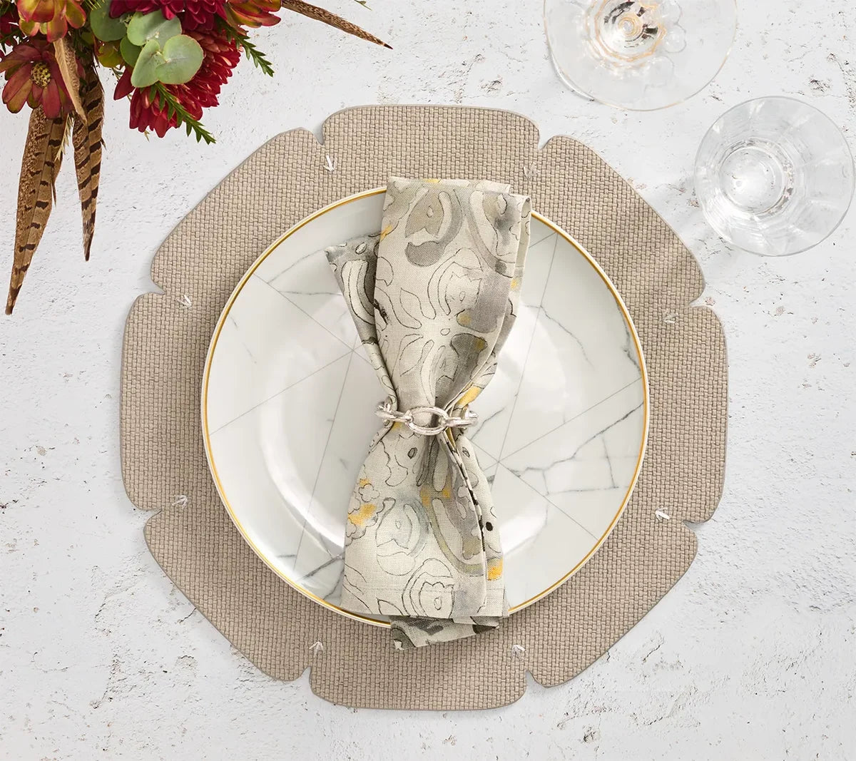 A grey Divot Placemat from Kim Seybert’s set of four features a scalloped edge and is paired with a marble-patterned plate, a cloth napkin tied with string, and two clear wine glasses on a white textured surface, beautifully complemented by a red and green floral arrangement in the corner—an elegant addition to your collection of dining room accessories.
