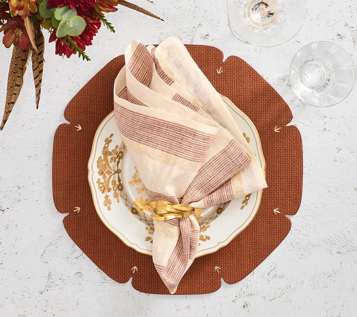 A gold and white plate with a folded striped napkin and gold napkin ring rests on the Kim Seybert Divot Placemat Set of 4 in Brown, adding an elegant touch to the dining table. Two glasses and a flower placed nearby on a white surface complete the modern yet inviting table setting.