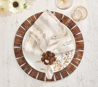A white plate with a wood-patterned rim sits on the Kim Seybert Halo Placemat in brown and white atop a white table. A folded napkin adorned with a bronze flower ring rests on the plate, beside two glasses of champagne and white flowers, creating an elegant table setting.