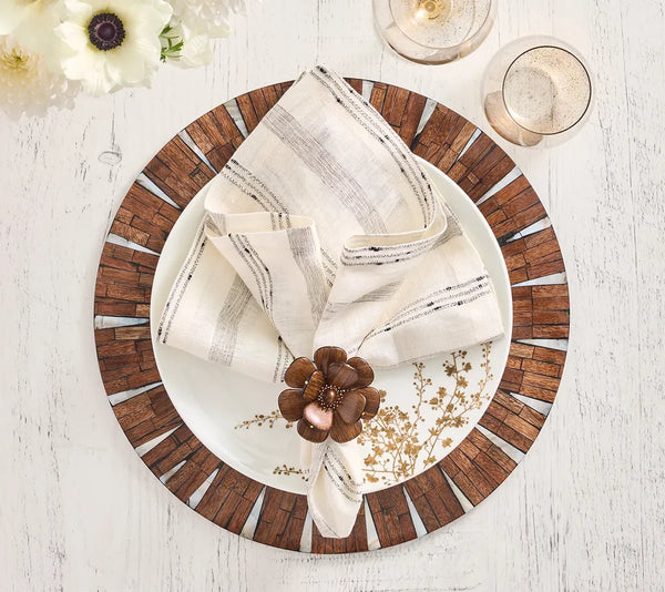 A white plate with a wood-patterned rim sits on the Kim Seybert Halo Placemat in brown and white atop a white table. A folded napkin adorned with a bronze flower ring rests on the plate, beside two glasses of champagne and white flowers, creating an elegant table setting.