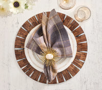 A plaid napkin with a gold starburst napkin ring rests on a white plate with gold dots, set atop the Kim Seybert Halo Placemat in brown and white. Two glasses and a white flower are nearby on a rustic white table.