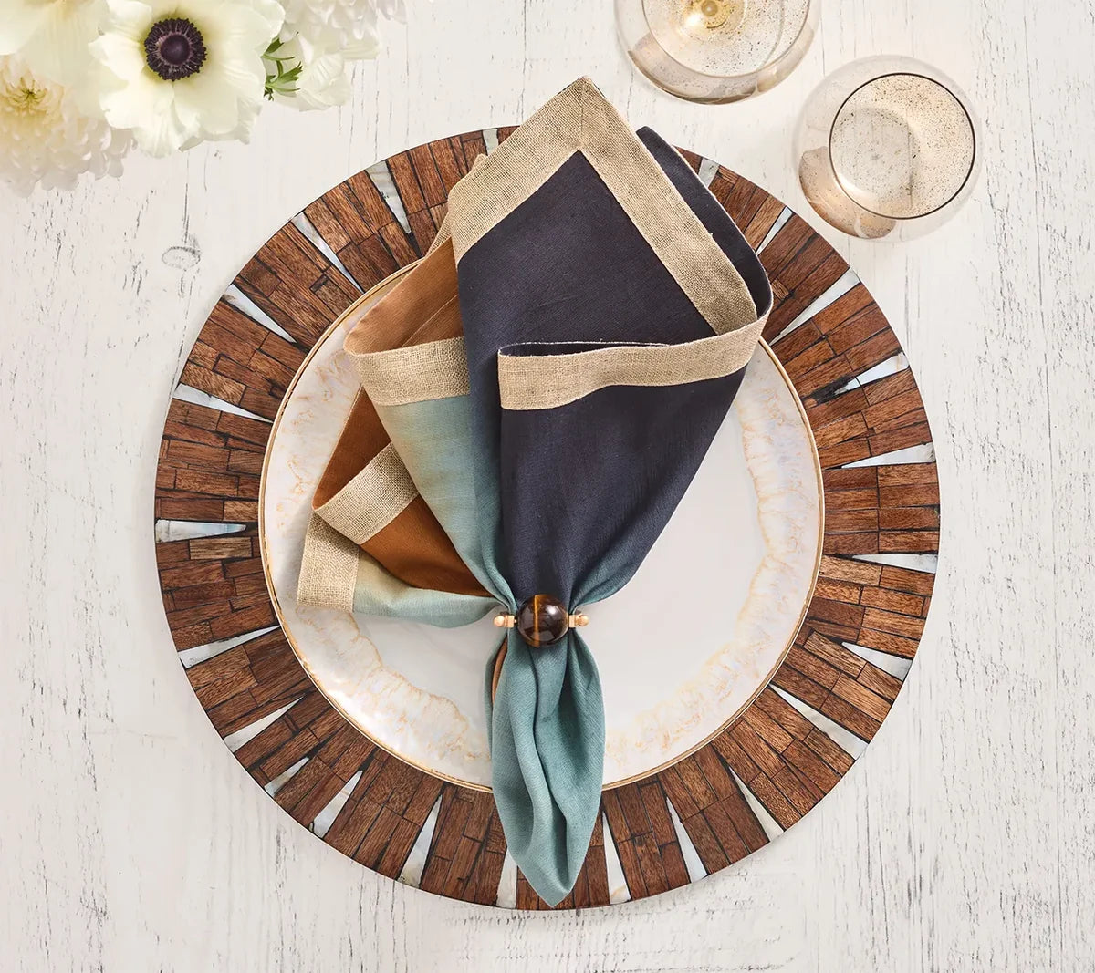 A decorative plate sits atop the Kim Seybert Halo Placemat in brown and white, layered over a wooden-patterned charger. A napkin in shades of blue, brown, and teal is neatly held by a ring. Two glasses and white flowers accompany the setting on a rustic white table.