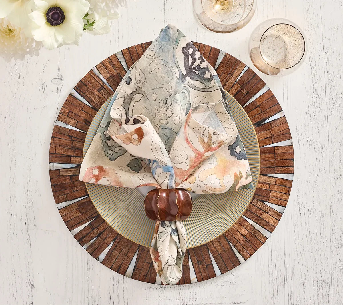 A patterned napkin with a wooden ring rests on a plate set atop the Kim Seybert Halo Placemat in brown and white, displayed on a white table. Two glasses and white flowers are placed nearby.