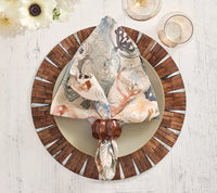 A patterned napkin with a wooden ring rests on a plate set atop the Kim Seybert Halo Placemat in brown and white, displayed on a white table. Two glasses and white flowers are placed nearby.