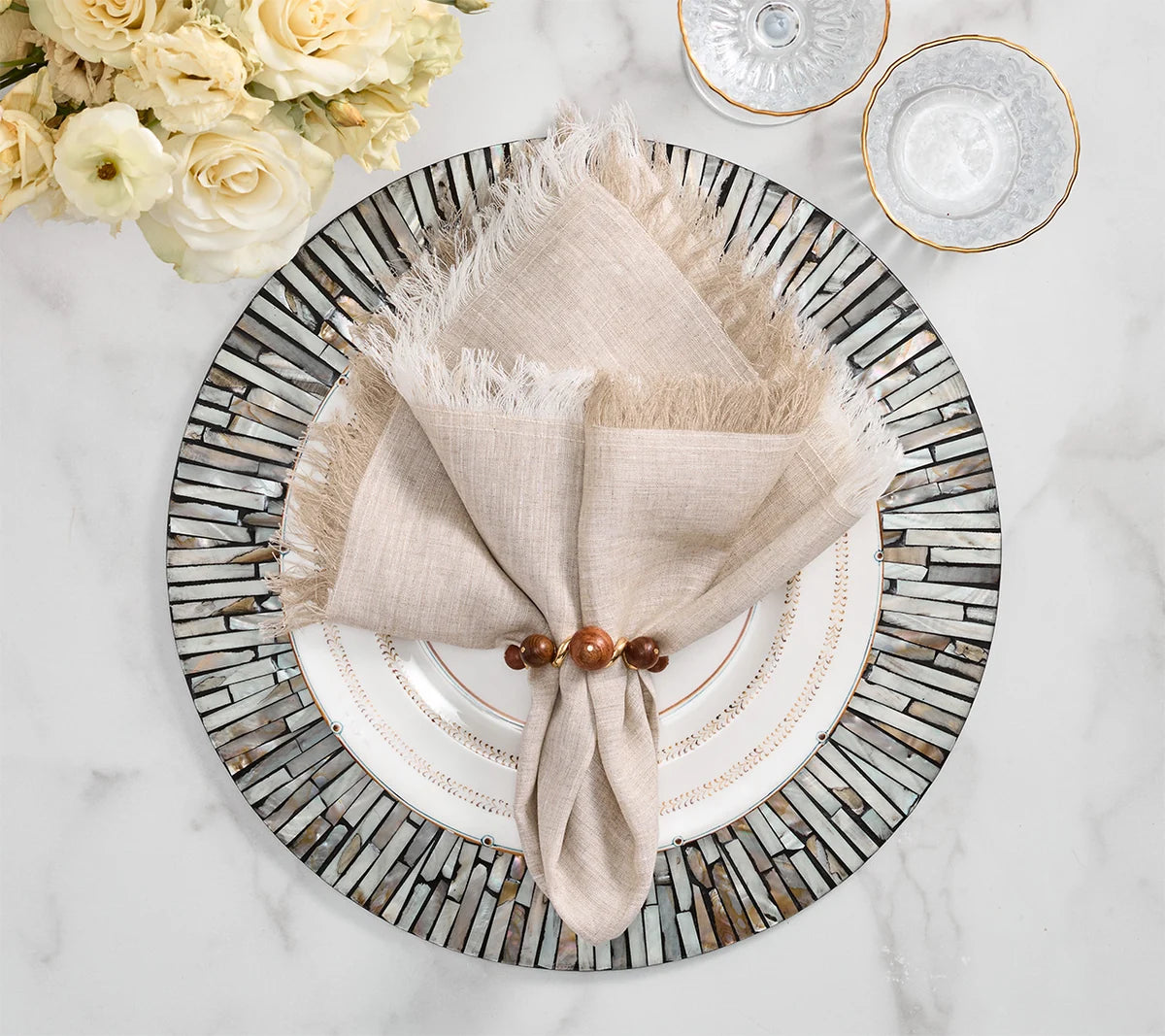 Overhead view of a table setting featuring the Kim Seybert Chambray Fringe Napkin Set of 4 in a wooden ring atop a decorative plate, arranged next to flowers and glassware on a marble surface for an inviting farmhouse-inspired decor touch.