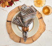A Windsor Napkin from Kim Seybert in a multicolored striped linen is paired with a modern napkin ring and placed on a white plate with a gold rim, set atop a wooden slice charger. Nearby, two amber glasses and autumn flowers in neutral tones are arranged on a marble surface.